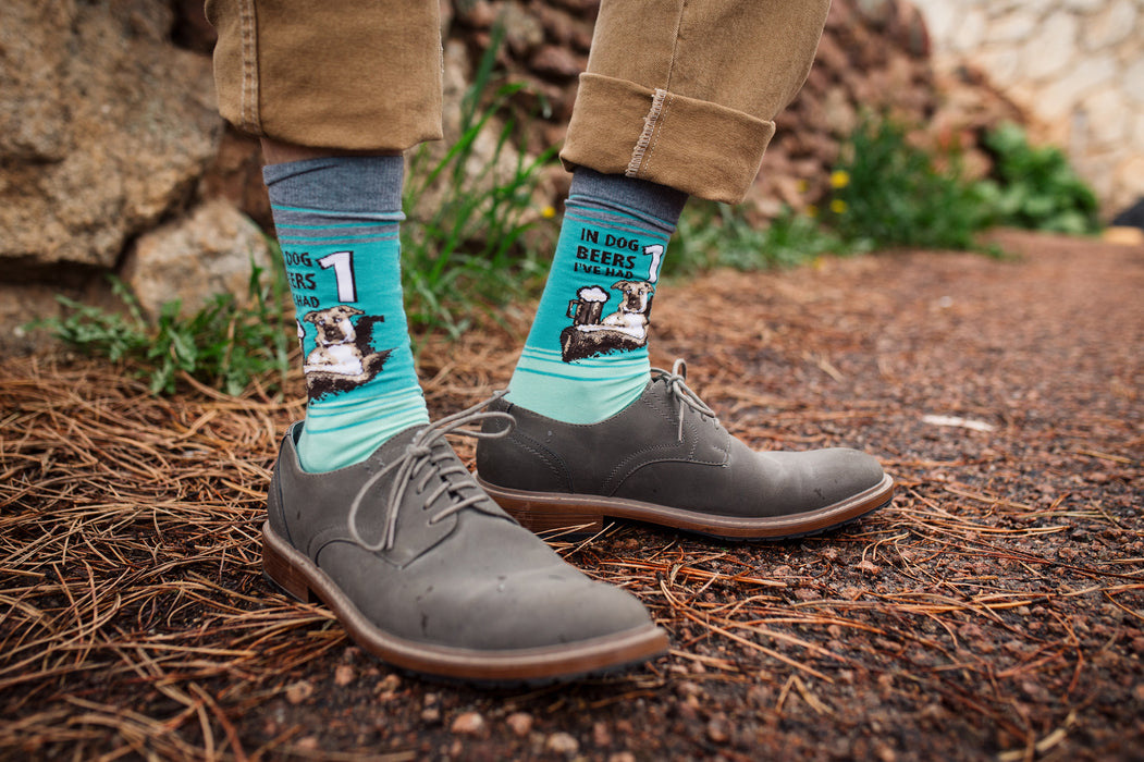 A person is sitting on a rock wall with their feet in brown shoes and blue socks with a pattern of cartoon dogs drinking beer. The person is wearing khaki pants and holding an open can of beer in their right hand.