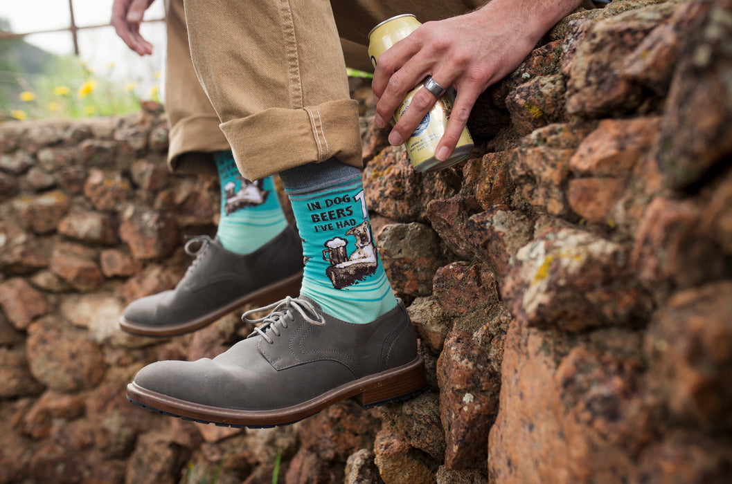 A person is sitting on a rock wall with their feet in brown shoes and blue socks with a pattern of cartoon dogs drinking beer. The person is wearing khaki pants and holding an open can of beer in their right hand.