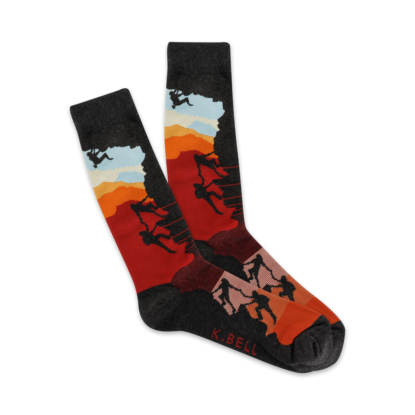 A pair of crew socks showcasing silhouetted climbers ascending a mountain against a vibrant sunset backdrop. The socks are mainly dark grey, red, and orange.