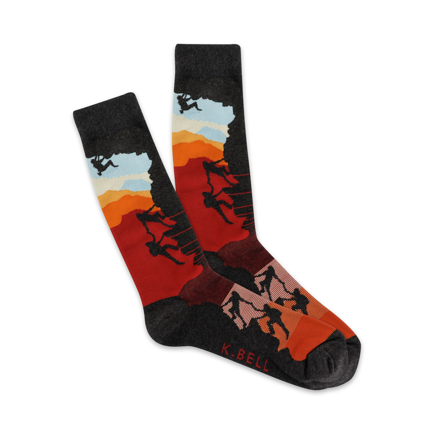 A pair of crew socks showcasing silhouetted climbers ascending a mountain against a vibrant sunset backdrop. The socks are mainly dark grey, red, and orange.