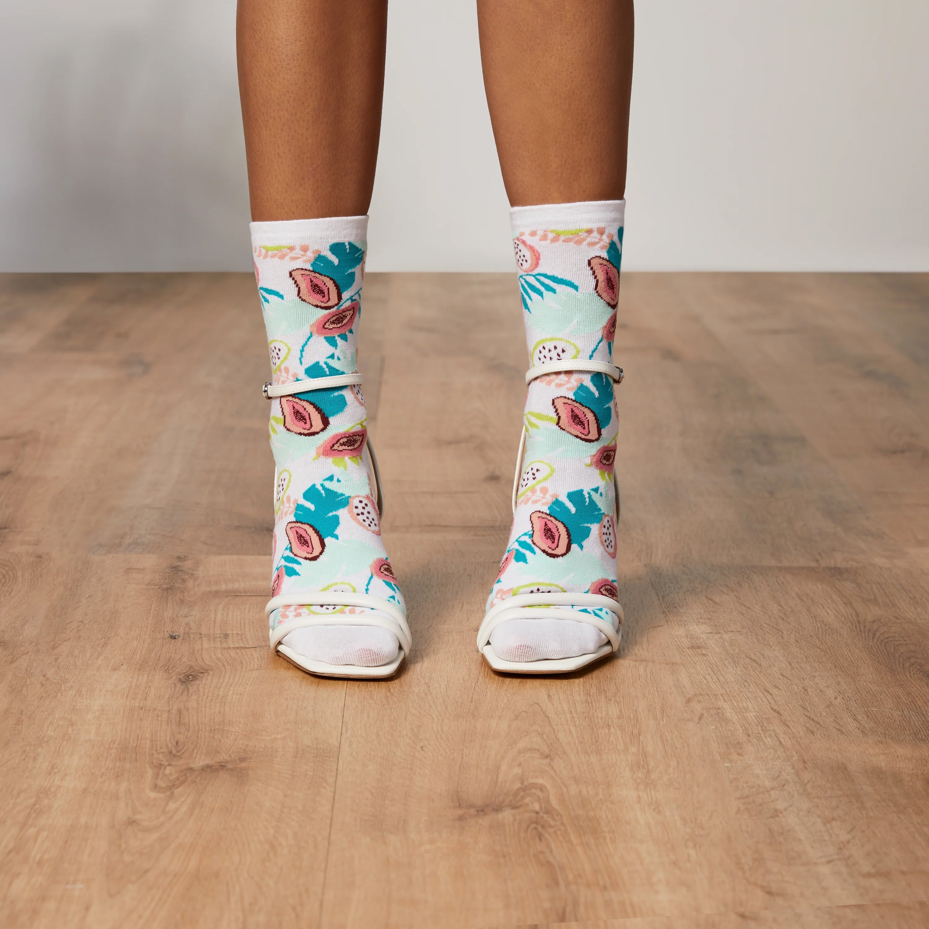 Lifestyle shot: Model's legs wearing Tropical Floral socks with white heels on a wood floor. [lf]