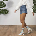 Lifestyle shot: Model wearing Tropical Floral socks with denim shorts and white heels; against a backdrop of large green leaves. [lf]