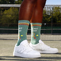 Lifestyle shot of a person wearing the 'I'd Hit That' socks with white sneakers on a tennis court. [lf]