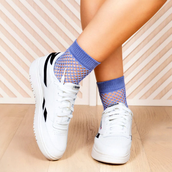 A person is wearing white sneakers with blue socks that have a fishnet-like pattern. The person is standing on a light-colored wooden floor.
