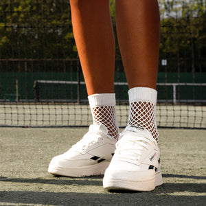 A young woman is seated on a chair with her legs crossed at the knee. She is wearing a black dress, white socks, and white sneakers with black trim. The sneakers are Reebok brand.