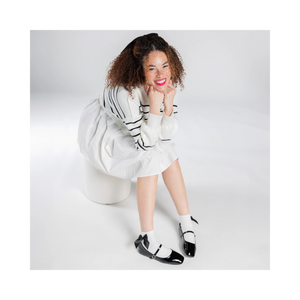 Overhead shot of a person wearing white ankle socks with a black bow and black patent leather shoes;  They're sitting down, smiling