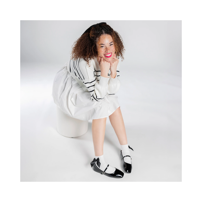 Overhead shot of a person wearing white ankle socks with a black bow and black patent leather shoes;  They're sitting down, smiling