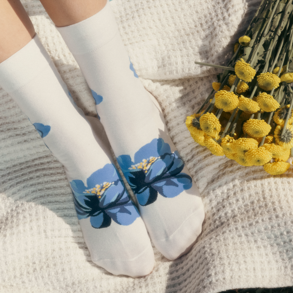 [lf] Lifestyle shot of The Big Bloom socks on a woman's feet, resting on a white knit blanket next to a bouquet of yellow flowers.