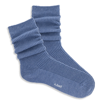 Pair of ribbed, slouchy crew socks in a dusty blue color, shown on a black background. 'k.bell' logo visible on sole.