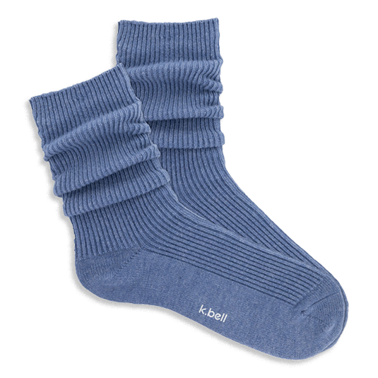 Pair of ribbed, slouchy crew socks in a dusty blue color, shown on a black background. 'k.bell' logo visible on sole.