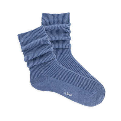 Pair of ribbed, slouchy crew socks in a dusty blue color, shown on a black background. 'k.bell' logo visible on sole.