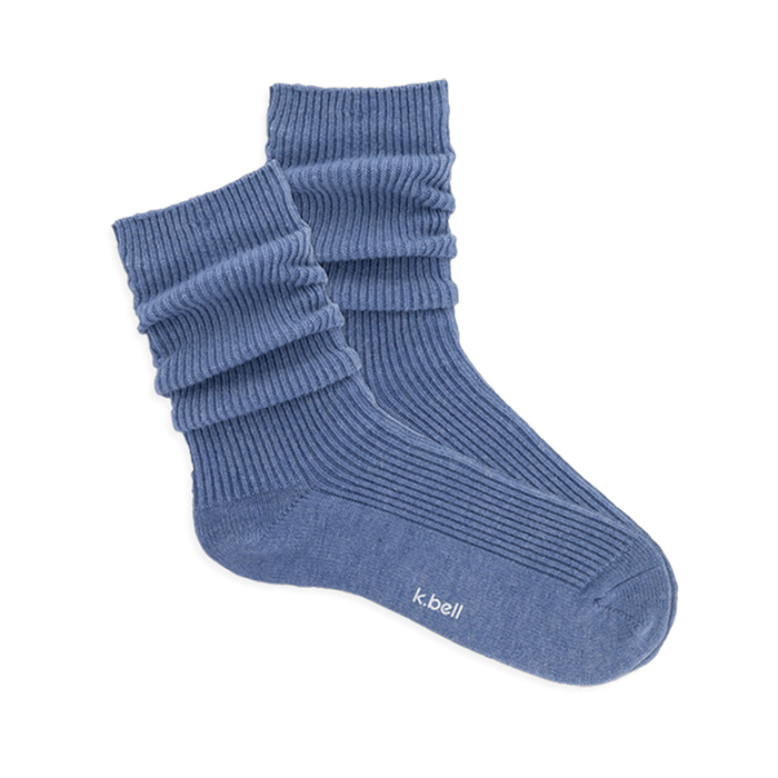 Pair of ribbed, slouchy crew socks in a dusty blue color, shown on a black background. 'k.bell' logo visible on sole.