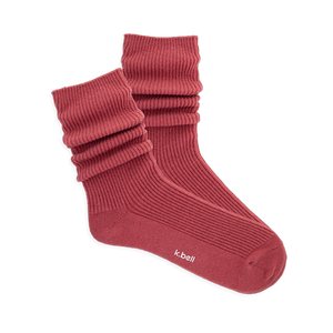 Pair of ribbed, slouchy crew socks in a muted red-orange color, shown on a black background. 'k.bell' logo visible on sole.