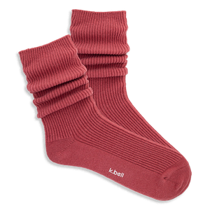 Pair of ribbed, slouchy crew socks in a muted red-orange color, shown on a black background. 'k.bell' logo visible on sole.