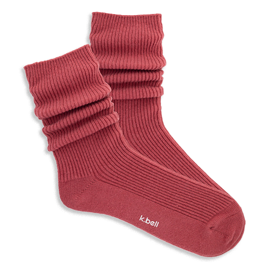 Pair of ribbed, slouchy crew socks in a muted red-orange color, shown on a black background. 'k.bell' logo visible on sole.