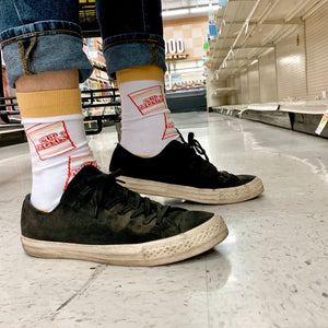 A person's feet in black sneakers and white socks with red and yellow Cup Noodles design. The person is standing in an empty grocery store aisle.