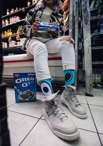 A person is sitting on a grocery store shelf with a box of Oreo cereal. The person is wearing white pants, gray sneakers, and socks with cartoon eyes on them.