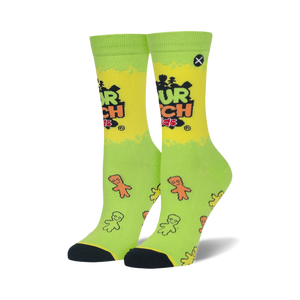 A pair of crew socks, lime green and yellow, with the Sour Patch Kids logo and scattered candy figures. Black toes and heels.