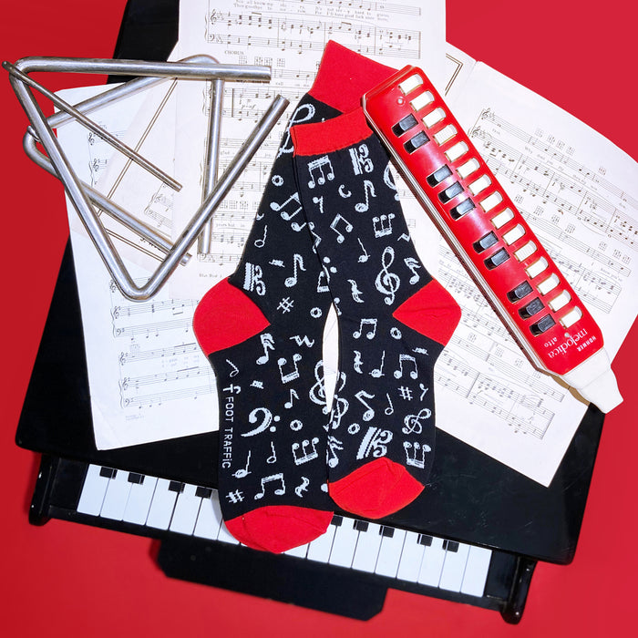 A pair of black socks with red toes and musical notes printed all over. The socks are laying on top of open sheet music with a red background. There is a silver triangle and a red and white harmonica also laying on the sheet music. The piano keys are visible underneath the sheet music.