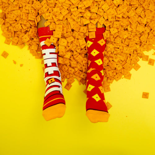 Lifestyle product shot: Feet in Cheez-It socks submerged in a pile of Cheez-It crackers against a yellow background. [lf]
