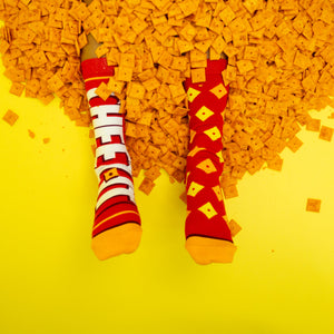 A pair of red and orange patterned socks is shown buried up to the ankles in a pile of small square crackers against a yellow background.