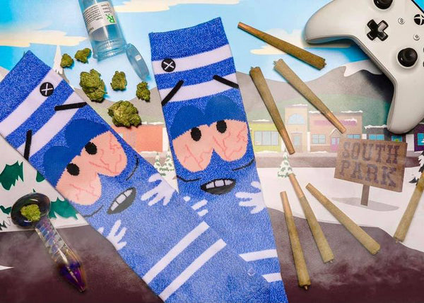 Lifestyle flat lay photo showing Towelie socks, marijuana paraphernalia, an Xbox controller and South Park themed background. [lf]