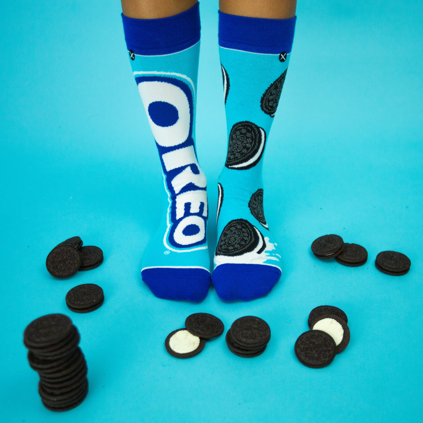 Lifestyle photo showing a person's feet wearing the Oreo socks surrounded by actual Oreo cookies on a blue background. [lf]