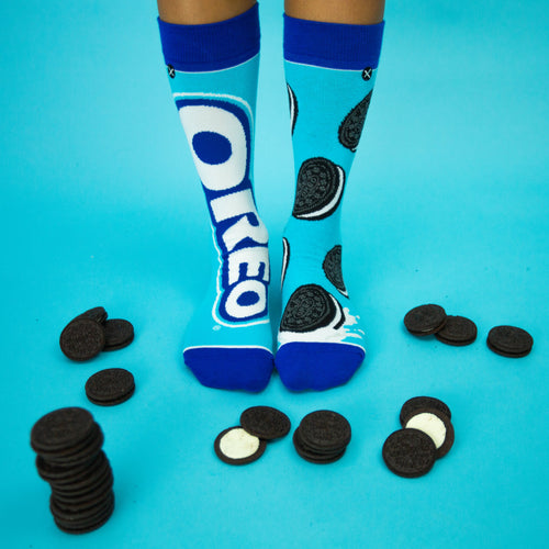 Lifestyle photo showing a person's feet wearing the Oreo socks surrounded by actual Oreo cookies on a blue background. [lf]