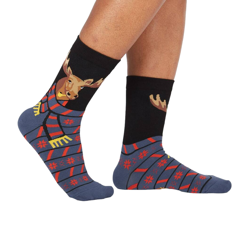 Lifestyle shot of a person's legs wearing the 'All Bundled Up' socks. The socks feature a moose wearing a scarf and snowflake pattern.
