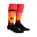 Pair of knee-high socks; red, orange, yellow, purple, and black. Design features an alien spaceship abducting a cow in a desert landscape at sunset.