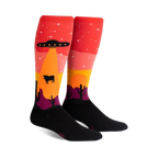 Pair of knee-high socks; red, orange, yellow, purple, and black. Design features an alien spaceship abducting a cow in a desert landscape at sunset.