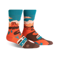 A pair of crew socks showcasing a Monument Valley scene. The socks are orange, teal, and brown, depicting a camper van driving along a desert road with cacti and rock formations.