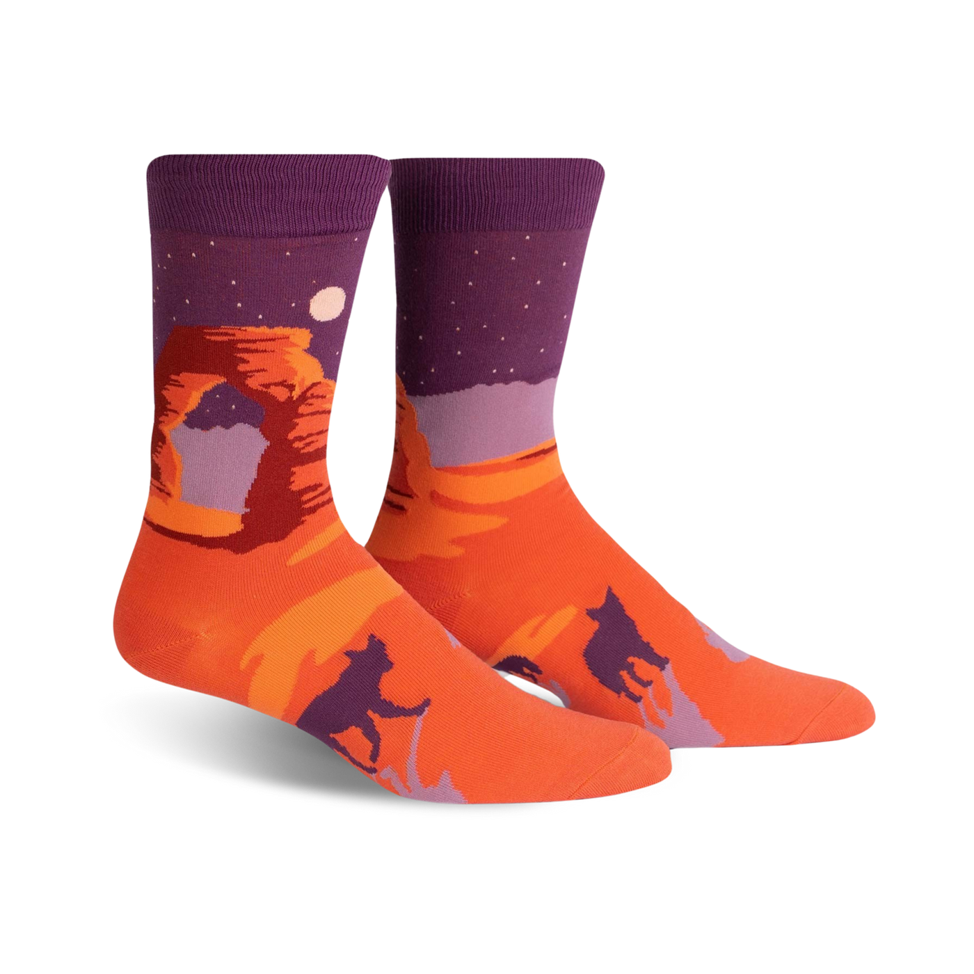 Crew socks in orange and purple, showcasing Delicate Arch at sunset under a starry night sky.  Silhouetted figures of coyotes or dogs are also featured.
