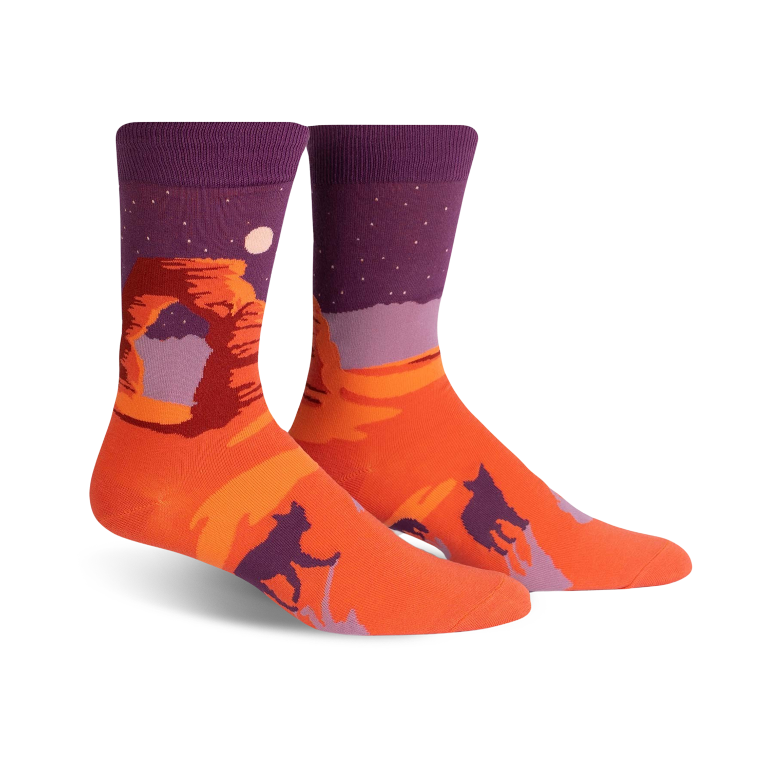 Crew socks in orange and purple, showcasing Delicate Arch at sunset under a starry night sky.  Silhouetted figures of coyotes or dogs are also featured.