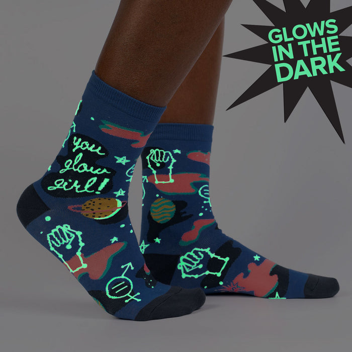 A pair of blue socks with a pattern of planets, stars, and other celestial bodies. The socks are pulled up on a person's legs, and the top of the socks is folded down to show the design. The socks are described as glowing in the dark.