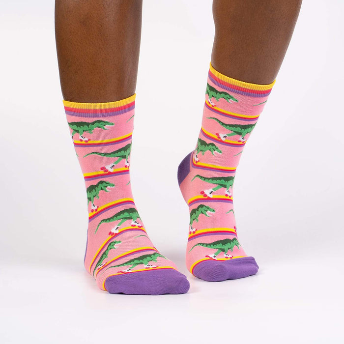 A pair of pink socks with a pattern of roller-skating dinosaurs wearing green dinosaur costumes. The socks have purple toes and heels with yellow and light blue stripes at the top.