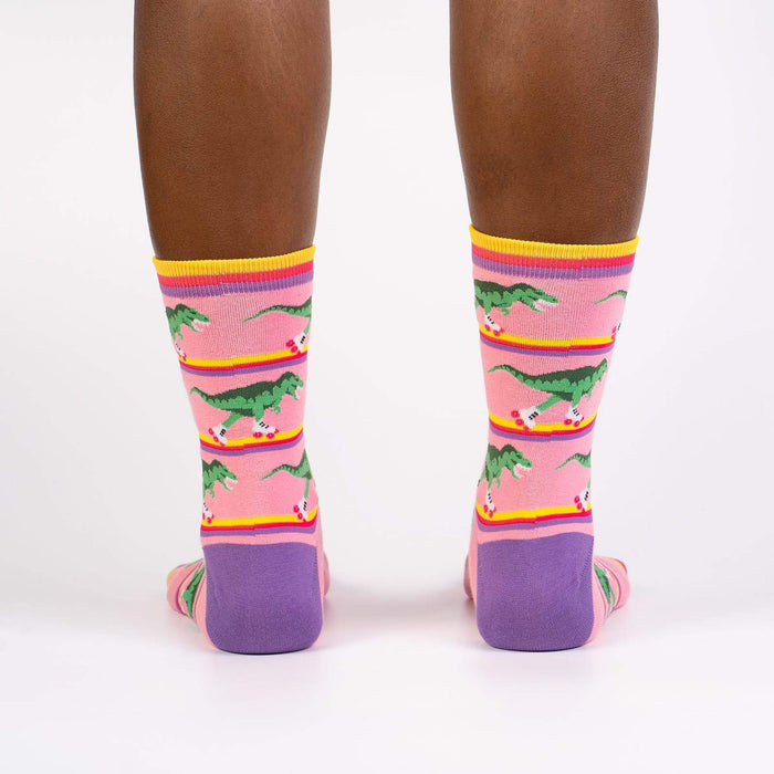 A pair of pink socks with a pattern of roller-skating dinosaurs wearing green dinosaur costumes. The socks have purple toes and heels with yellow and light blue stripes at the top.