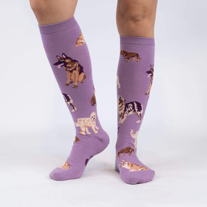 A pair of purple knee-high socks with a pattern of cartoon dogs on them. The dogs are of various breeds, including corgis, pugs, bulldogs, and beagles.