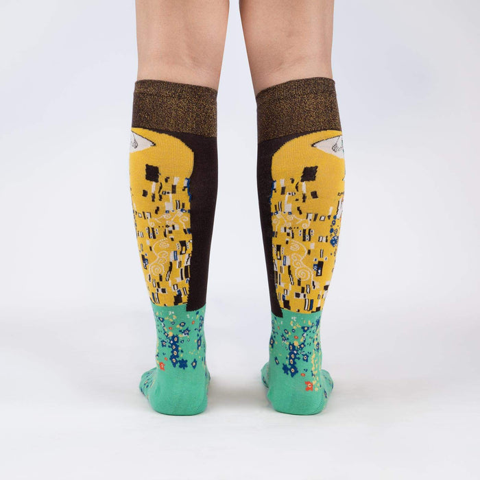 A pair of legs is shown from the back wearing a pair of knee-high socks with a Gustav Klimt-inspired pattern.