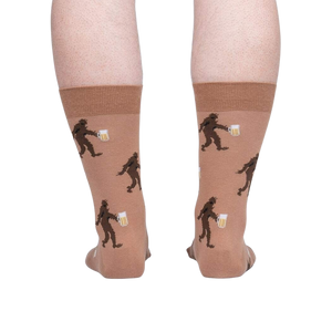 A pair of brown crew socks with a pattern of cartoonish Bigfoot creatures holding beers.