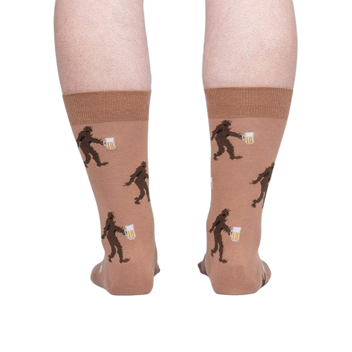 A pair of brown crew socks with a pattern of cartoonish Bigfoot creatures holding beers.