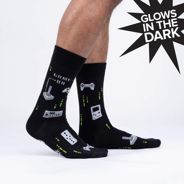 A pair of black socks with a pattern of 8-bit video game controllers and systems. The socks are described as glowing in the dark.