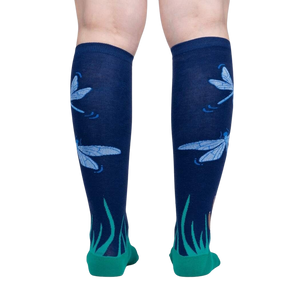 A pair of legs is shown from the back wearing a pair of dark blue knee socks with a pattern of blue and green dragonflies and green grass at the cuff.