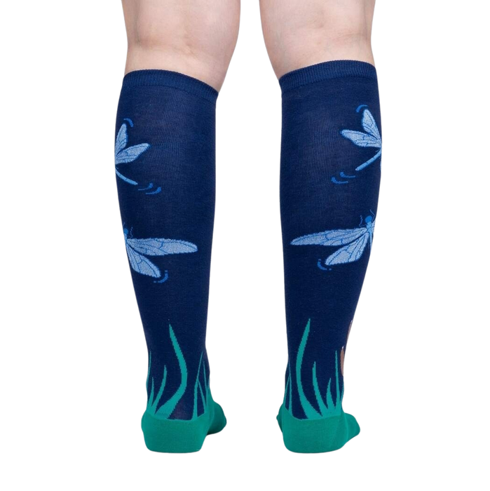 A pair of legs is shown from the back wearing a pair of dark blue knee socks with a pattern of blue and green dragonflies and green grass at the cuff.