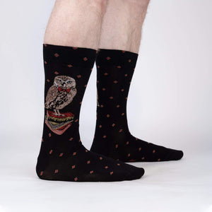 A pair of black socks with a pattern of brown owls wearing red bow ties standing on a stack of books. The socks are shown on the back of a person's legs.