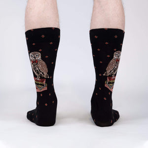 A pair of black socks with a pattern of brown owls wearing red bow ties standing on a stack of books. The socks are shown on the back of a person's legs.