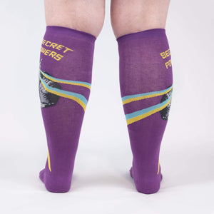 A pair of purple knee-high socks with a design of a narwhal swimming with a yellow and blue trail behind it. The word 