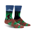 A pair of crew socks showcasing Bigfoot trekking through a forest landscape with a mountain in the background. The socks are predominantly blue, green, and brown, with a fern pattern at the bottom.