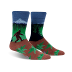 A pair of crew socks showcasing Bigfoot trekking through a forest landscape with a mountain in the background. The socks are predominantly blue, green, and brown, with a fern pattern at the bottom.