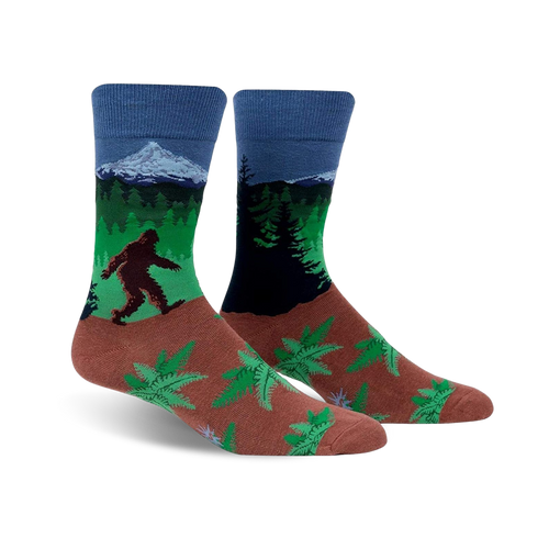 A pair of crew socks showcasing Bigfoot trekking through a forest landscape with a mountain in the background. The socks are predominantly blue, green, and brown, with a fern pattern at the bottom.